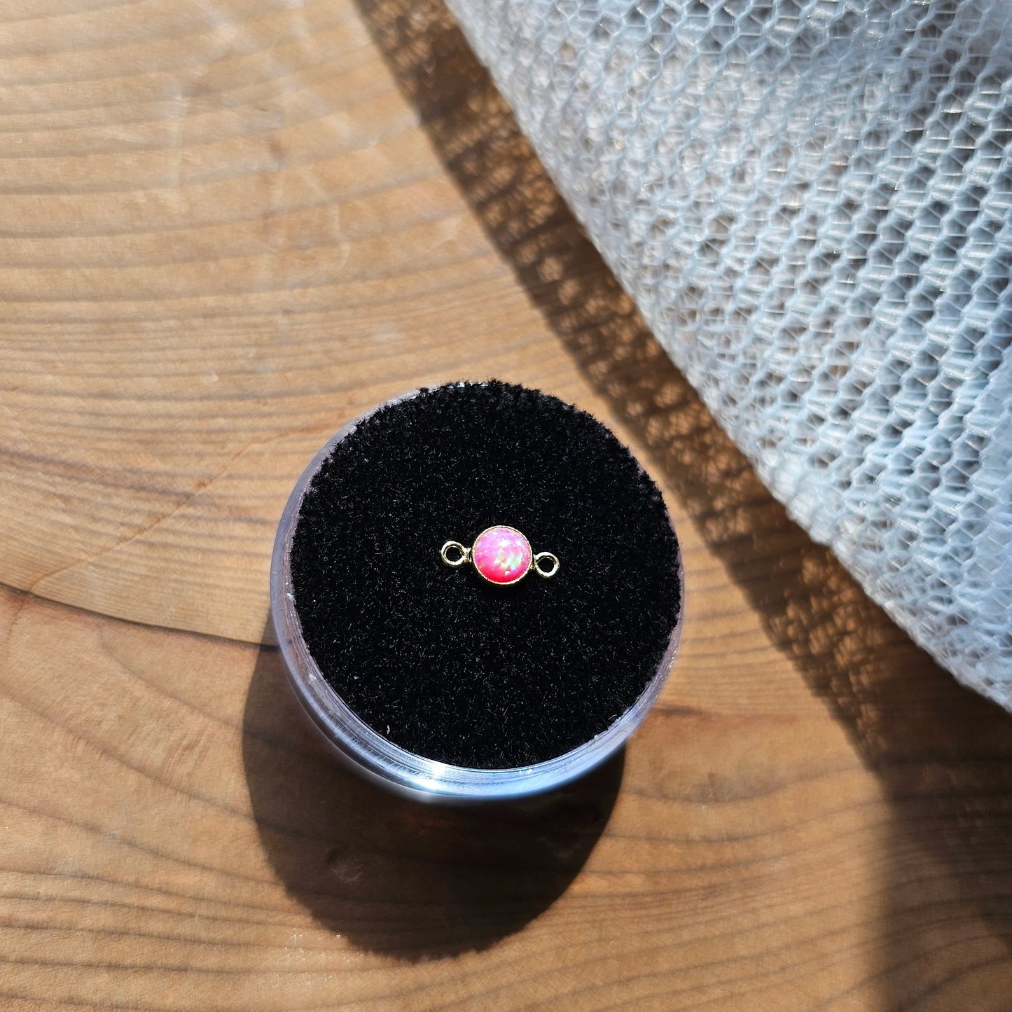 Iridescent charm connector for permanent jewelry in pink opal in the sunlight on a wood stand
